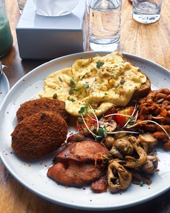 A plate of diverse breakfast items including scrambled eggs topped with greens, two crispy brown patties, roasted mushrooms, grilled bacon, and a serving of baked beans with tomatoes. The meal is set on a wooden table alongside a tissue box and a glass of water.