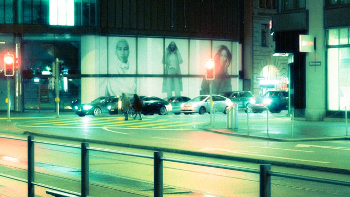 A bustling urban street at night with cars waiting at a traffic light. Large illuminated advertisements featuring people are displayed on a building in the background. Pedestrians are crossing the zebra crossing, and the scene is bathed in a mix of green and blue artificial lighting.