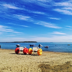 Colorful pedal boats with large wheels rest on a sandy beach with the sea in the background. A clear blue sky with white clouds stretches above, and a small island is visible in the distance.