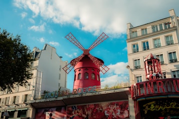 A red windmill structure stands prominently against a bright blue sky, surrounded by classic French architecture. The windmill is part of a famous entertainment venue, with intricate designs and a vibrant presence. Trees and traditional Parisian buildings flank the scene, creating a blend of nature and urban charm.