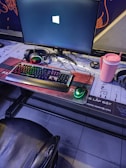 A gaming setup featuring a large monitor displaying a Windows logo, a colorful RGB keyboard, a pair of headphones, a gaming mouse with green lighting, and a pink tumbler with a straw. The setup is on a desk with a patterned mouse pad, situated next to a black padded chair.