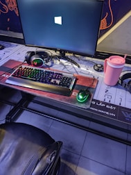 A gaming setup featuring a large monitor displaying a Windows logo, a colorful RGB keyboard, a pair of headphones, a gaming mouse with green lighting, and a pink tumbler with a straw. The setup is on a desk with a patterned mouse pad, situated next to a black padded chair.