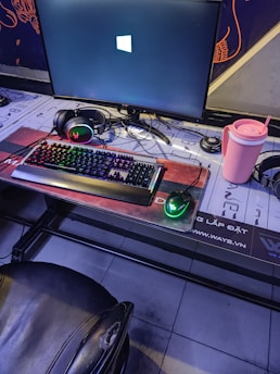 A gaming setup featuring a large monitor displaying a Windows logo, a colorful RGB keyboard, a pair of headphones, a gaming mouse with green lighting, and a pink tumbler with a straw. The setup is on a desk with a patterned mouse pad, situated next to a black padded chair.
