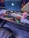 A gaming setup featuring a large monitor displaying a Windows logo, a colorful RGB keyboard, a pair of headphones, a gaming mouse with green lighting, and a pink tumbler with a straw. The setup is on a desk with a patterned mouse pad, situated next to a black padded chair.