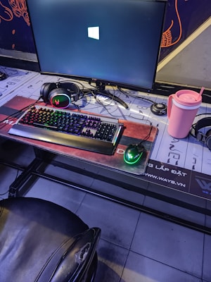 A gaming setup featuring a large monitor displaying a Windows logo, a colorful RGB keyboard, a pair of headphones, a gaming mouse with green lighting, and a pink tumbler with a straw. The setup is on a desk with a patterned mouse pad, situated next to a black padded chair.