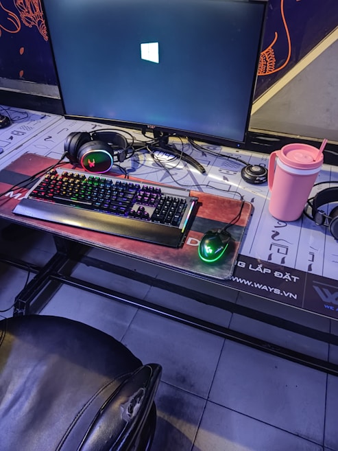 A gaming setup featuring a large monitor displaying a Windows logo, a colorful RGB keyboard, a pair of headphones, a gaming mouse with green lighting, and a pink tumbler with a straw. The setup is on a desk with a patterned mouse pad, situated next to a black padded chair.