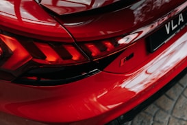 A red sports car tail light with sleek design elements and reflective surfaces. The image focuses on a prominent RS badge near the rear of the vehicle, with intricate light patterns that create a dynamic visual impression. The surface reflects surrounding light and patterns, enhancing the car's aesthetic appeal.