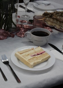 A piece of layered cake with a cream filling and red fruit jam on a white plate. Next to the plate is a white cup filled with dark coffee, labeled 'Cappuccino.' The table is decorated with metallic pink leaves and a clear glass containing a candle. Some other plates and utensils are visible, along with a vase holding a bouquet of flowers.