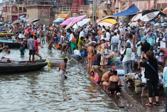 A bustling riverside scene at a ghats where a large number of people are gathered. Some individuals are bathing in the river, while others are engaged in various activities like washing clothes, praying, or taking photos. Colorful umbrellas and clothes add vibrancy to the setting, and boats are docked nearby.