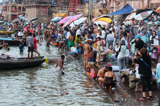 A bustling riverside scene at a ghats where a large number of people are gathered. Some individuals are bathing in the river, while others are engaged in various activities like washing clothes, praying, or taking photos. Colorful umbrellas and clothes add vibrancy to the setting, and boats are docked nearby.
