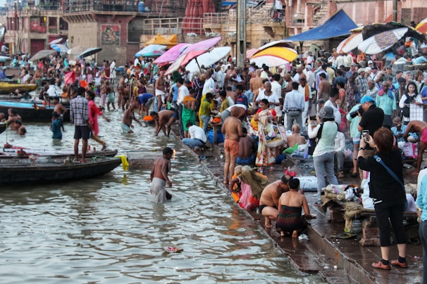 A bustling riverside scene at a ghats where a large number of people are gathered. Some individuals are bathing in the river, while others are engaged in various activities like washing clothes, praying, or taking photos. Colorful umbrellas and clothes add vibrancy to the setting, and boats are docked nearby.
