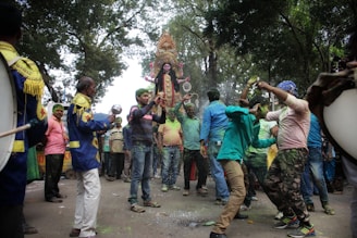 A vibrant street procession with a group of people enthusiastically dancing and celebrating around an ornate statue of a goddess. The scene is lively, with colorful clothing and musical instruments, and takes place under a canopy of trees.