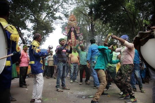 A vibrant street procession with a group of people enthusiastically dancing and celebrating around an ornate statue of a goddess. The scene is lively, with colorful clothing and musical instruments, and takes place under a canopy of trees.