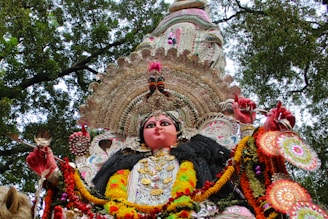 A richly adorned idol featuring vibrant colors, intricate designs, and traditional attire, set against a backdrop of tree branches and foliage. The idol is embellished with detailed patterns, jewelry, and floral garlands.