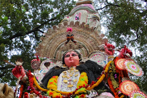 A richly adorned idol featuring vibrant colors, intricate designs, and traditional attire, set against a backdrop of tree branches and foliage. The idol is embellished with detailed patterns, jewelry, and floral garlands.