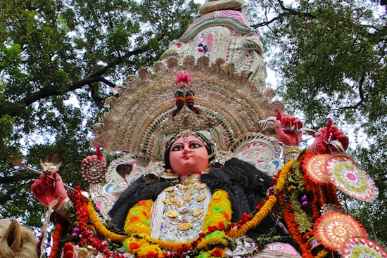 A richly adorned idol featuring vibrant colors, intricate designs, and traditional attire, set against a backdrop of tree branches and foliage. The idol is embellished with detailed patterns, jewelry, and floral garlands.