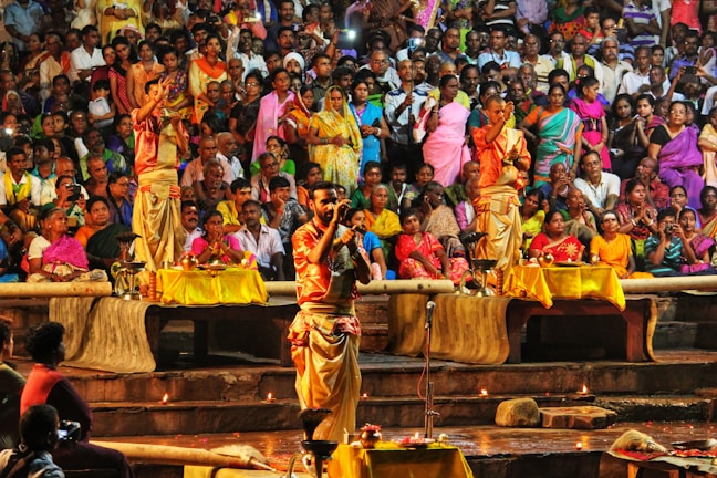 A large group of people gathered in a public space, with a focus on three individuals performing a ritual. The performers are dressed in traditional attire and are surrounded by decorated tables with offerings. Many of the onlookers are seated in colorful clothing, some taking photos or recording the event. The atmosphere is vibrant and lively, indicating a cultural or religious ceremony.