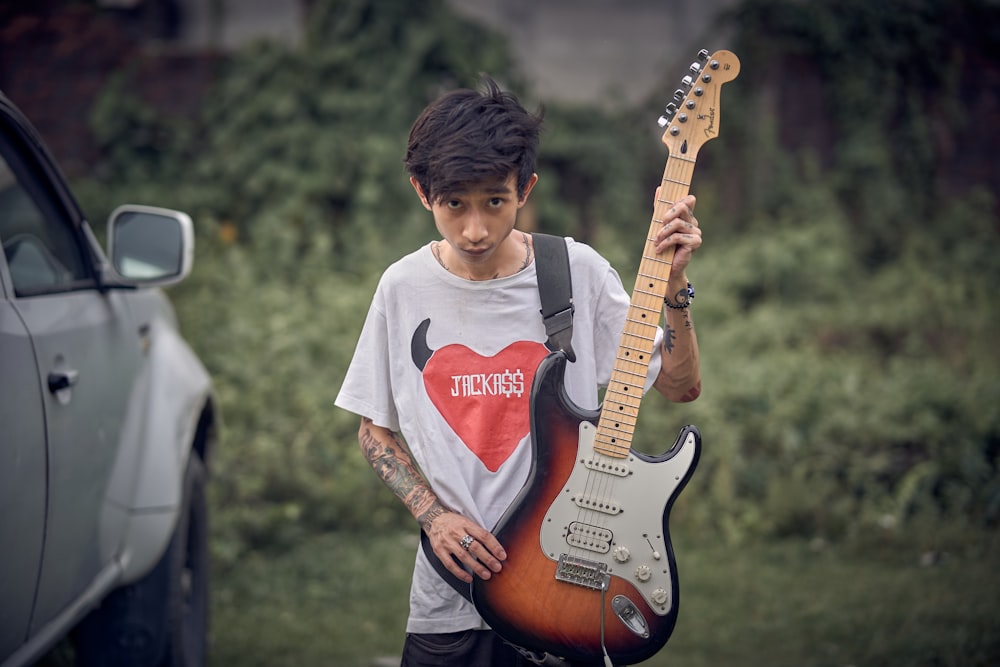 a young man holding a guitar in front of a car