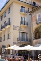 A multi-story building with 'Hotel Dell'Angelo' and 'Ristorante' signs, featuring several balconies and large windows. The ground floor hosts a restaurant with outdoor seating under large umbrellas. Patrons are seen dining, and the adjacent building showcases decorative architectural details.