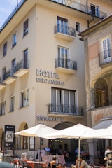 A multi-story building with 'Hotel Dell'Angelo' and 'Ristorante' signs, featuring several balconies and large windows. The ground floor hosts a restaurant with outdoor seating under large umbrellas. Patrons are seen dining, and the adjacent building showcases decorative architectural details.