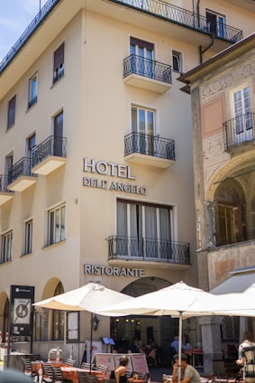 A multi-story building with 'Hotel Dell'Angelo' and 'Ristorante' signs, featuring several balconies and large windows. The ground floor hosts a restaurant with outdoor seating under large umbrellas. Patrons are seen dining, and the adjacent building showcases decorative architectural details.