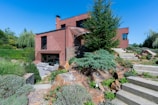A modern brick house with geometric architecture features amidst a landscaped garden. The scene includes green lawns, various shrubs, rocks, and stone steps leading up to the entrance. Outdoor furniture is positioned on a patio area, offering a serene and inviting atmosphere.