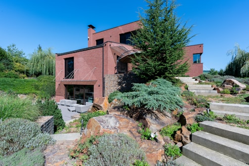 A modern brick house with geometric architecture features amidst a landscaped garden. The scene includes green lawns, various shrubs, rocks, and stone steps leading up to the entrance. Outdoor furniture is positioned on a patio area, offering a serene and inviting atmosphere.
