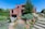 A modern brick house with geometric architecture features amidst a landscaped garden. The scene includes green lawns, various shrubs, rocks, and stone steps leading up to the entrance. Outdoor furniture is positioned on a patio area, offering a serene and inviting atmosphere.