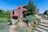A modern brick house with geometric architecture features amidst a landscaped garden. The scene includes green lawns, various shrubs, rocks, and stone steps leading up to the entrance. Outdoor furniture is positioned on a patio area, offering a serene and inviting atmosphere.