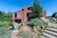 A modern brick house with geometric architecture features amidst a landscaped garden. The scene includes green lawns, various shrubs, rocks, and stone steps leading up to the entrance. Outdoor furniture is positioned on a patio area, offering a serene and inviting atmosphere.