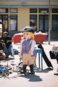 A large toy figure resembling a person is prominently displayed outdoors amidst various market stalls. The figure is wearing a helmet and has a friendly appearance, standing next to a tricycle and a blue table. People and various items are visible in the background, creating a busy, market-like atmosphere.