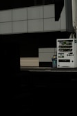 A vending machine stands on the right side of the image, next to a recycling bin. It's situated against the backdrop of a building with a grid-pattern facade. Most of the image is in shadow, giving a stark contrast between the lit vending machine and the dark foreground.