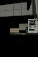 A vending machine stands on the right side of the image, next to a recycling bin. It's situated against the backdrop of a building with a grid-pattern facade. Most of the image is in shadow, giving a stark contrast between the lit vending machine and the dark foreground.