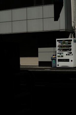 A vending machine stands on the right side of the image, next to a recycling bin. It's situated against the backdrop of a building with a grid-pattern facade. Most of the image is in shadow, giving a stark contrast between the lit vending machine and the dark foreground.