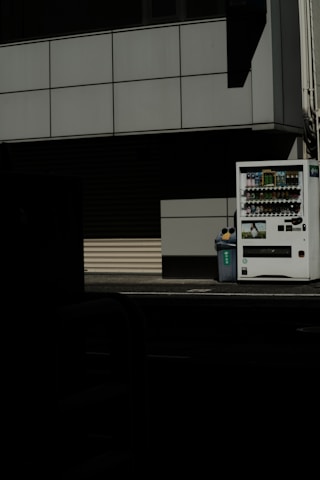 A vending machine stands on the right side of the image, next to a recycling bin. It's situated against the backdrop of a building with a grid-pattern facade. Most of the image is in shadow, giving a stark contrast between the lit vending machine and the dark foreground.