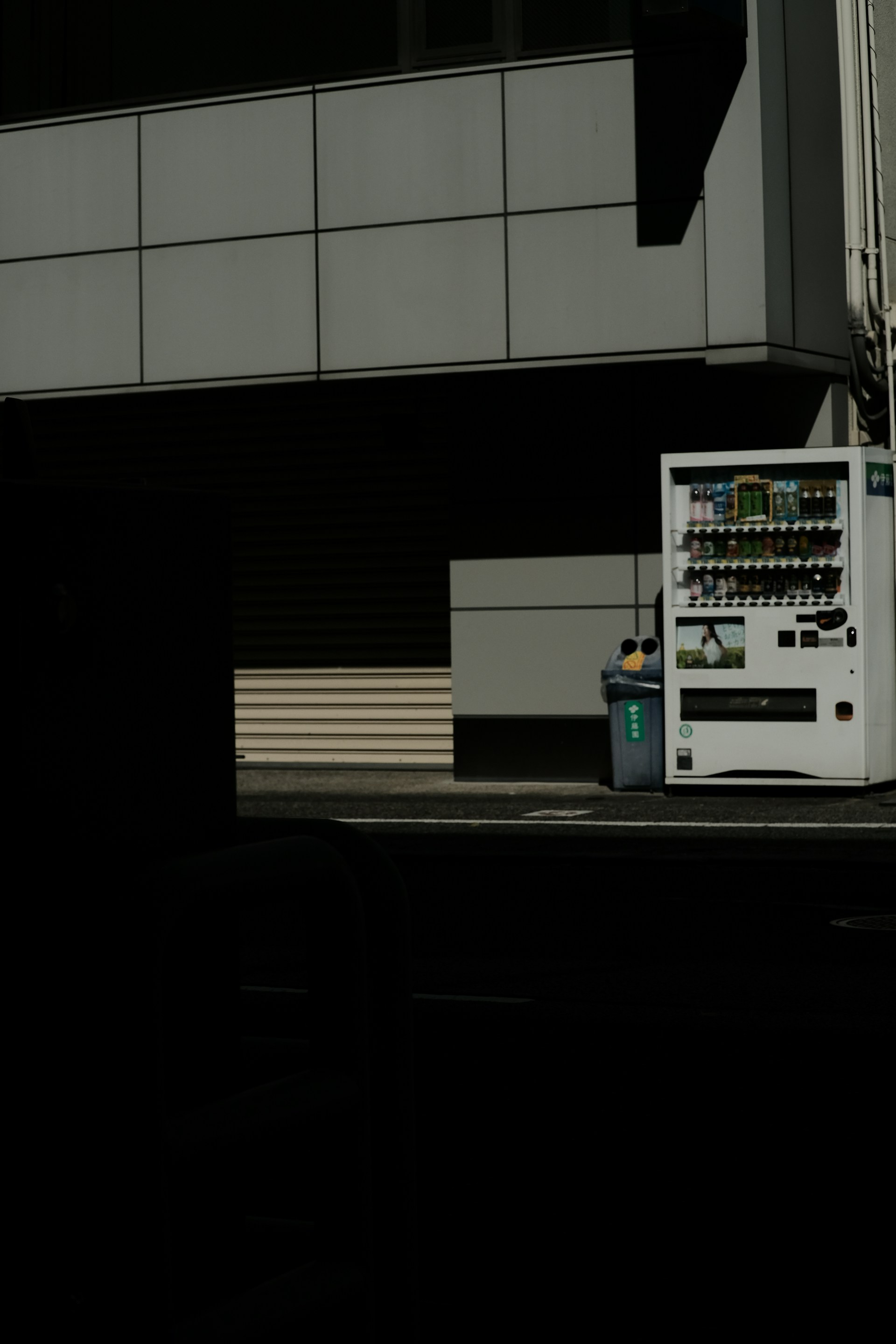 A vending machine stands on the right side of the image, next to a recycling bin. It's situated against the backdrop of a building with a grid-pattern facade. Most of the image is in shadow, giving a stark contrast between the lit vending machine and the dark foreground.