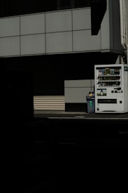 A vending machine stands on the right side of the image, next to a recycling bin. It's situated against the backdrop of a building with a grid-pattern facade. Most of the image is in shadow, giving a stark contrast between the lit vending machine and the dark foreground.