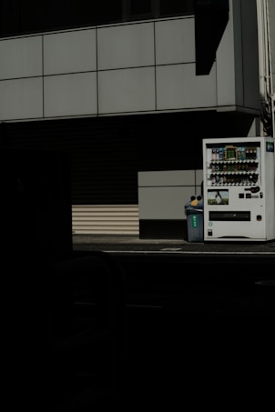 A vending machine stands on the right side of the image, next to a recycling bin. It's situated against the backdrop of a building with a grid-pattern facade. Most of the image is in shadow, giving a stark contrast between the lit vending machine and the dark foreground.