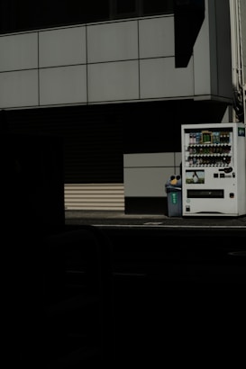 A vending machine stands on the right side of the image, next to a recycling bin. It's situated against the backdrop of a building with a grid-pattern facade. Most of the image is in shadow, giving a stark contrast between the lit vending machine and the dark foreground.