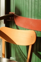 A wooden chair with a light brown finish is positioned in front of a green paneled wall, partially illuminated by sunlight. A horizontal red-brown railing is attached to the wall, creating strong shadows across the scene.