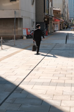 A person wearing a dark suit and face mask walks down a sunlit, paved pedestrian street. The street features buildings on both sides with various shopfronts and signage. Shadows are cast across the pavement, and the scene evokes a sense of quiet urban life.