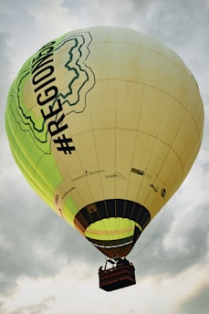 A large hot air balloon is floating in the sky, featuring a design with green contour lines and the text '#REGIONS' on its side. The balloon is mainly beige with some green and black accents. A basket is attached below, carrying a few passengers who are partially visible. The sky is overcast with gray clouds surrounding the balloon.