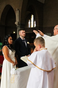 An indoor wedding ceremony with a bride in a white dress and a groom in a dark suit standing before a priest. The setting is a church with arched windows and brick walls. A child in a white robe stands nearby holding a book.