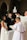 An indoor wedding ceremony with a bride in a white dress and a groom in a dark suit standing before a priest. The setting is a church with arched windows and brick walls. A child in a white robe stands nearby holding a book.