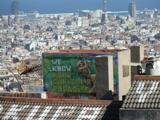 A densely packed urban area with a mix of modern and historic architecture under a clear blue sky. In the foreground, a rooftop is visible with graffiti that reads 'WE KNOW YOUR CAPITALIST PARADISE' in bold colors. A figure is painted next to the text.