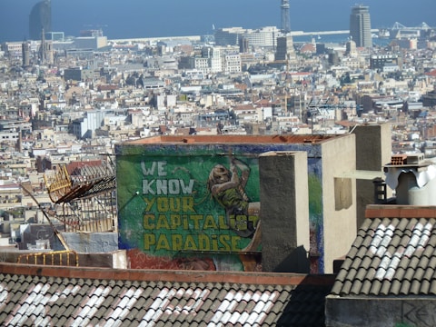 A densely packed urban area with a mix of modern and historic architecture under a clear blue sky. In the foreground, a rooftop is visible with graffiti that reads 'WE KNOW YOUR CAPITALIST PARADISE' in bold colors. A figure is painted next to the text.