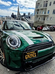 A green Mini Cooper with a California license plate is parked on a street. The city skyline is visible in the background, featuring iconic skyscrapers under a partly cloudy sky. Buildings and other parked cars are nearby.