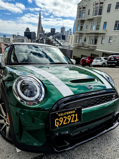 A green Mini Cooper with a California license plate is parked on a street. The city skyline is visible in the background, featuring iconic skyscrapers under a partly cloudy sky. Buildings and other parked cars are nearby.