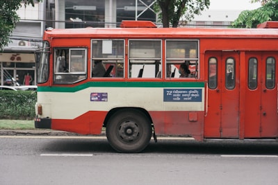 A red and cream bus parked on a street, with a green stripe running along its side. There are passengers visible through the windows, and a blue sign with text is displayed on the side of the bus. Trees and a building can be seen in the background.