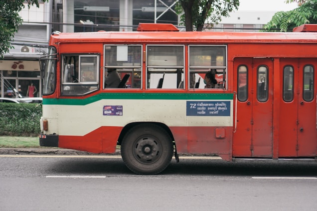 A red and cream bus parked on a street, with a green stripe running along its side. There are passengers visible through the windows, and a blue sign with text is displayed on the side of the bus. Trees and a building can be seen in the background.